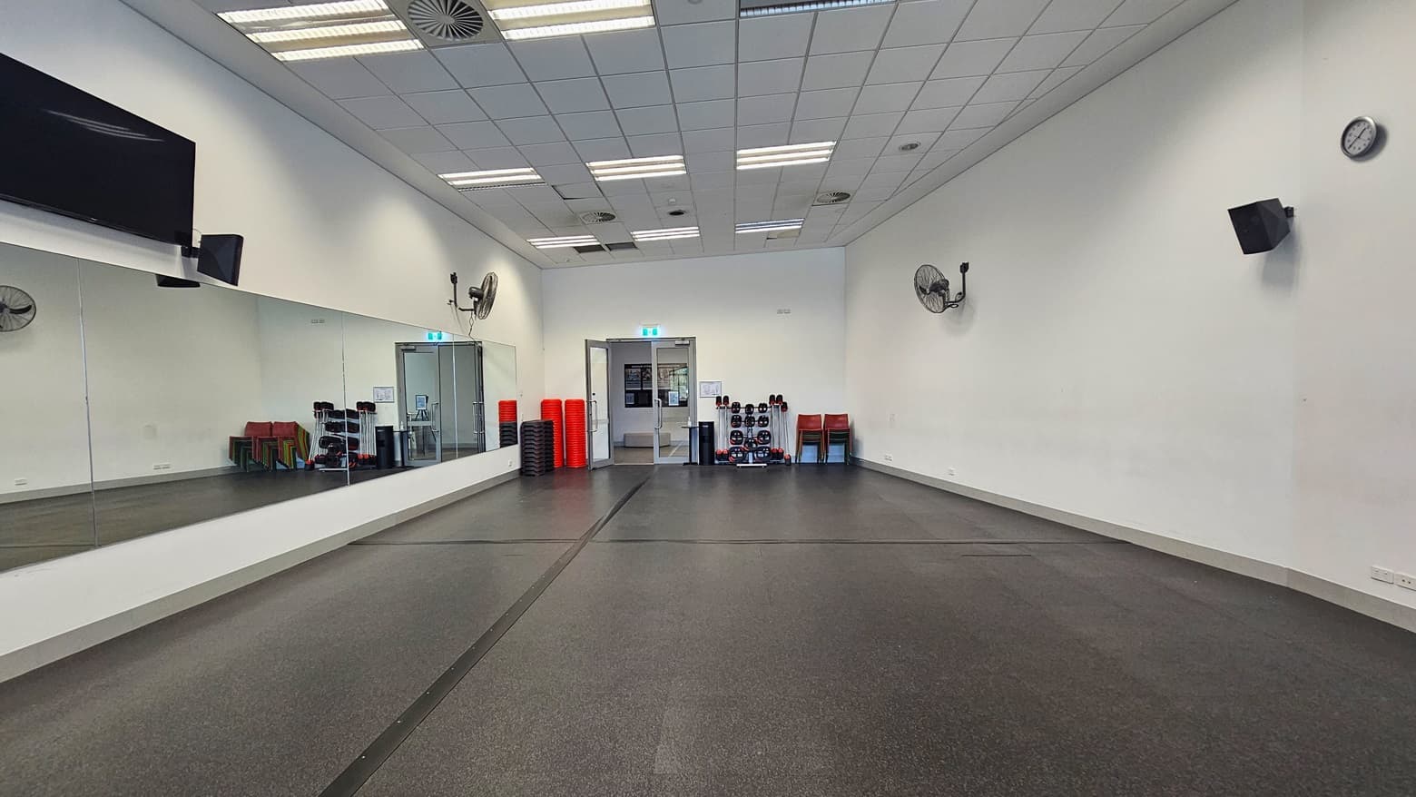 The fitness room is spacious, with black rubber tiles covering the floor. Mirrors line the left wall, while a flat-screen TV, wall fans, a clock, and audio speakers are affixed to the upper walls. In the middle, an inward-opening glass door is present. Fitness equipment is arranged near the door, alongside red plastic chairs. The ceiling and walls are consistently white.