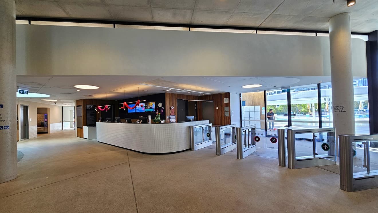 The reception desk is a white counter with a brick design. On the countertop and ceiling are computers and Christmas decorations. The reception wall has a dark wooden design. On the left, a lobby leads to other facilities. The floor is concrete, and the ceiling is white with LED lights. To the right, turnstiles marked for separate entry and exit are present, with the Aquatic facility also visible.