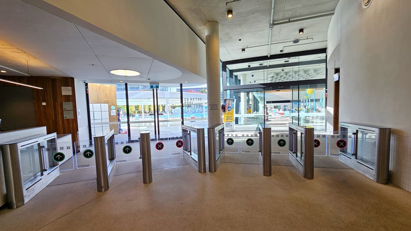 In the centre are glass turnstiles marked for separate entry and exit. Beyond the gates, another glass entrance door leads to the Aquatic Centre. On the left, a brown wall displays posters, while the right wall is white. The ceiling is white with pin lights and LED lights. The floor is constructed of concrete.