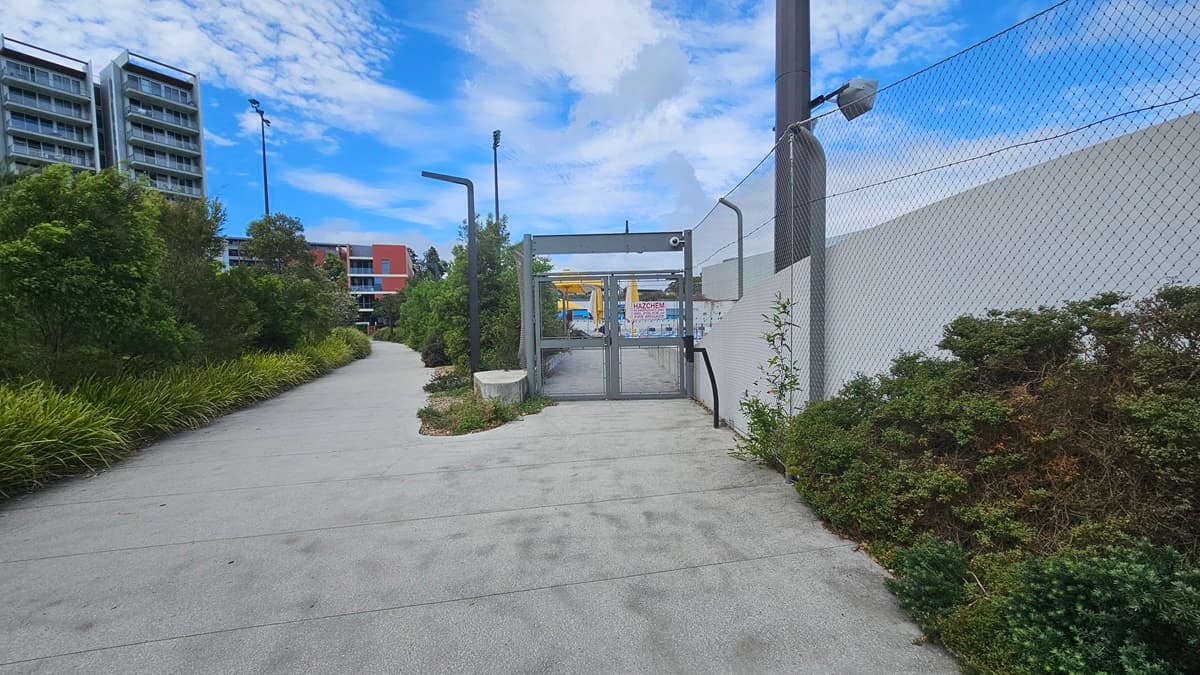 To the left, a tall commercial building, trees, and ornamental plants border the road. In the middle, a concrete path leads to a gate with a glass door. To the right, a white wall with a chain fence is present. The hillside section on the right is covered with wild grass and shrubs.