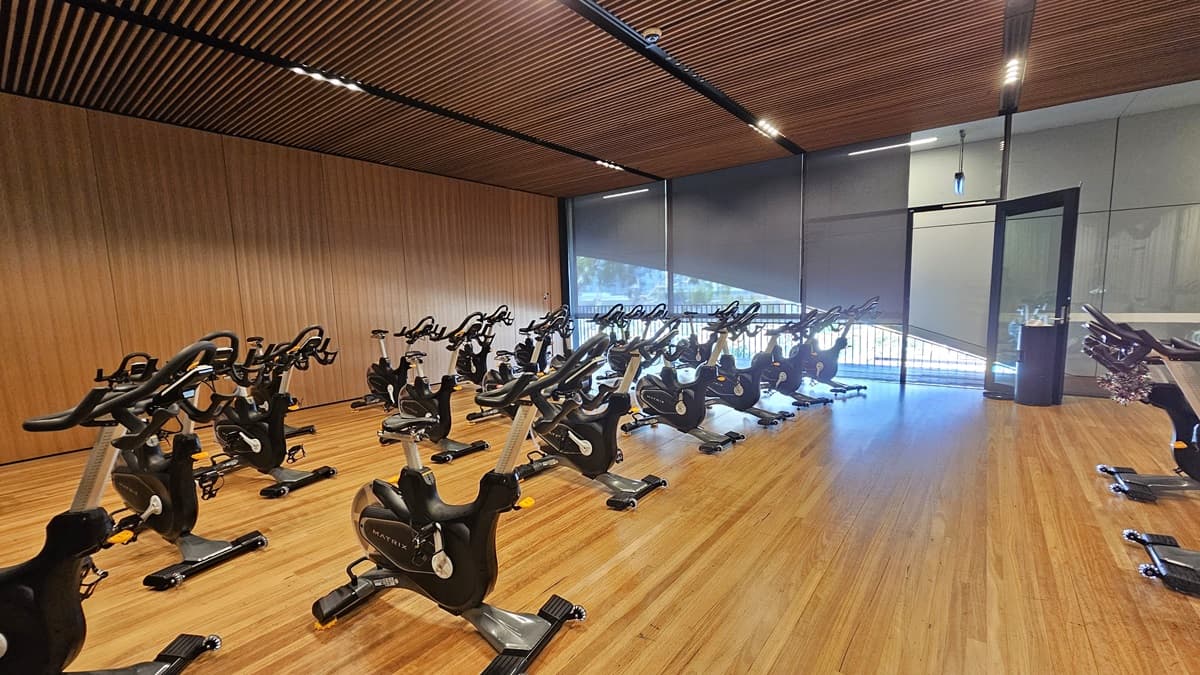 Spin machines are arranged in rows, filling the timber-floored room. An inward-opening glass door serves as an entry to this space. A black rubbish bin is placed near the door. The right wall is made of glass, and the left wall has a timber design with a wooden plank ceiling design equipped with LED lights.