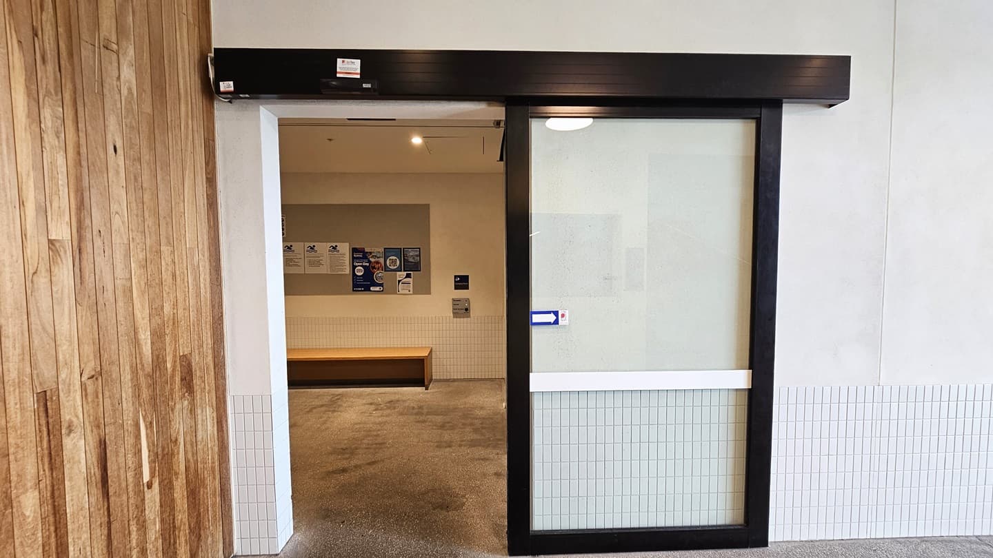 The entrance to the Hydrotherapy Pool is through an automatic glass sliding door. The left wall features a timber design, and the right wall is white concrete. Inside, the wall is equipped with safety banners and reminder signs. A wooden bench chair is placed against the wall.