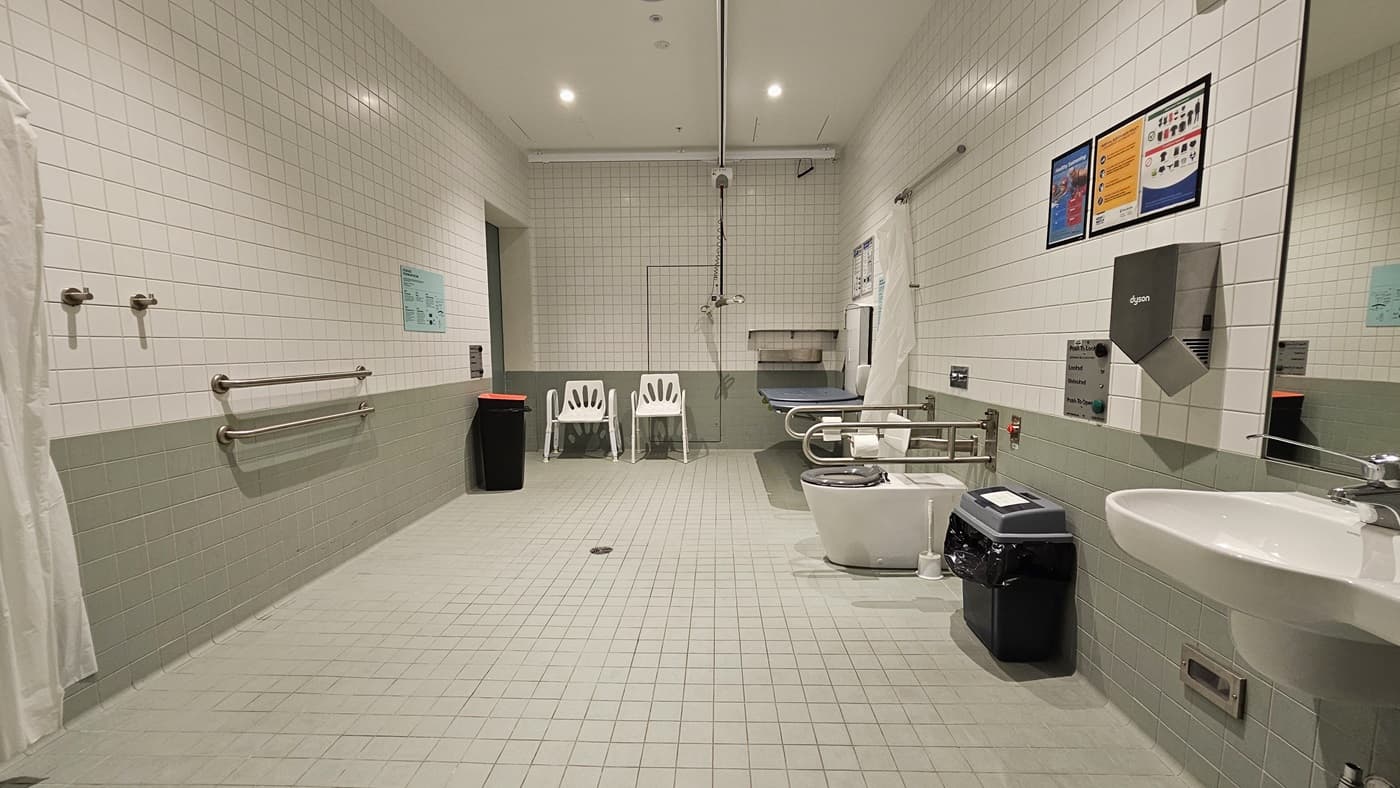On the right, there's a foldable change table with a ceiling hoist. Adjacent is the toilet bowl with hand grabs, rubbish bins, and access buttons. A wash basin, mirror, and another rubbish bin are on the right. The ceiling is all white, and the walls are white with green accents. To the right, the wooden stairs are visible through the glass wall.