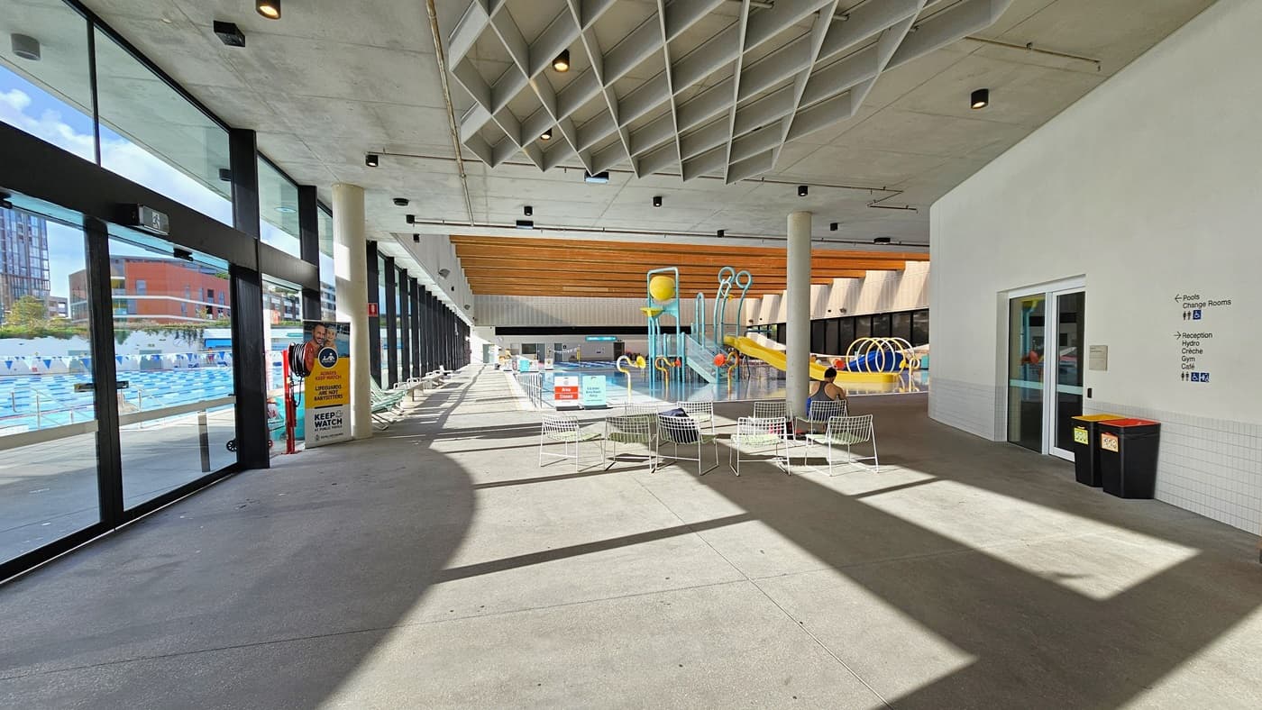 In the middle, movable white chairs with mesh designs are arranged. To the right, a white wall with signage for the accessible toilet is present. Rubbish bins are visible. Behind, the Aqua Play Pool features a yellow slide with fountains, tipping buckets, and water features. A retractable banner is positioned near the glass wall, and safety banners are placed on the pool deck. The ceiling is white with diamond-shaped wooden structures and wooden planks.