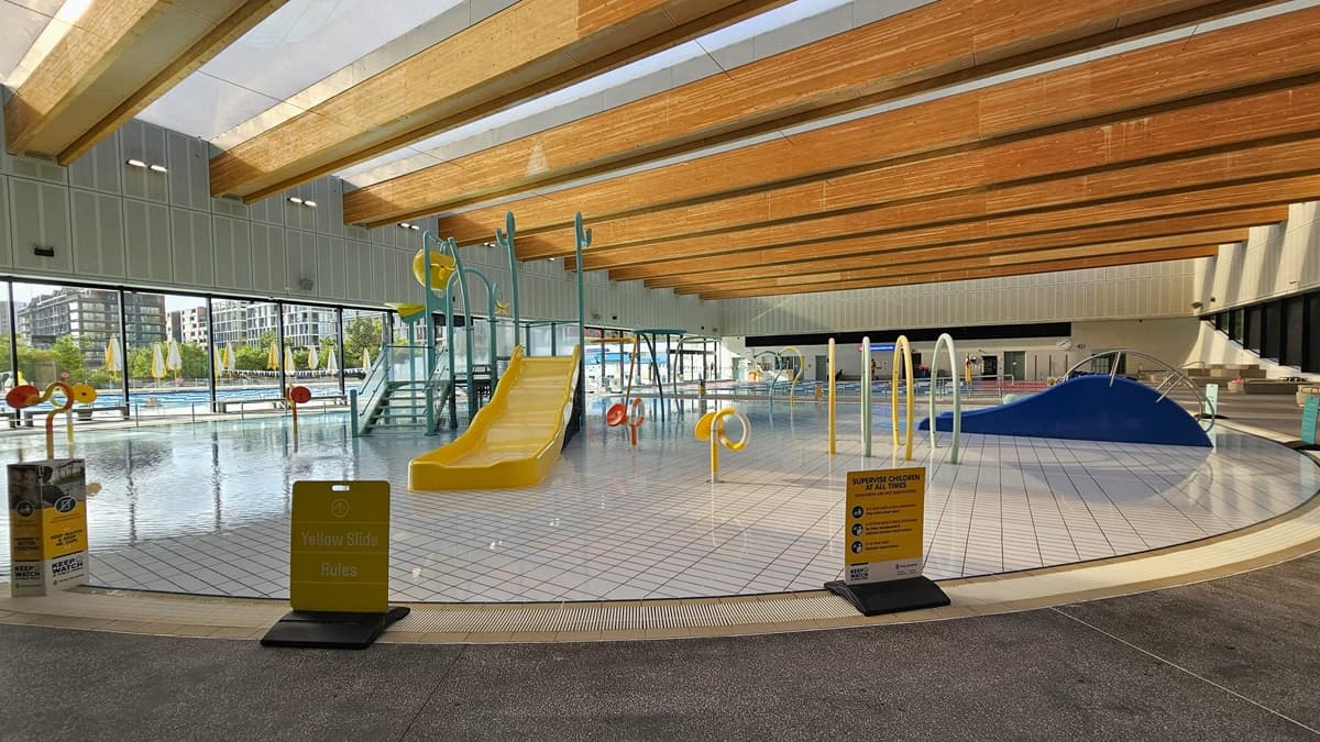 The Aqua Play Pool has colourful structures, a yellow slide with sprayers, tipping buckets, fountains, and other water features, including a wading area. An overflow grid surrounds the pool, and safety banners are placed on the pool deck. The ceiling features a wooden plank design with floodlights. The left wall has glass walls providing a view of the outdoor aquatic facility.