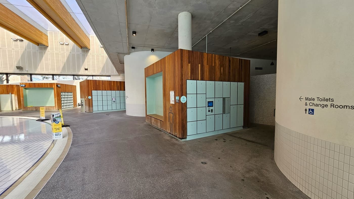To the right are Male and Female Toilets and Change Rooms in Cabana style. The bathroom features a dark brown wood panel and white tiles design. To the left is the Aqua Play Pool, equipped with an overflow grid and safety banners. The wall on the right is white with small brick accents. A sign stating "Accessible Bathroom" is written on the wall. The ceiling features a transparent white ceiling with timber accents.