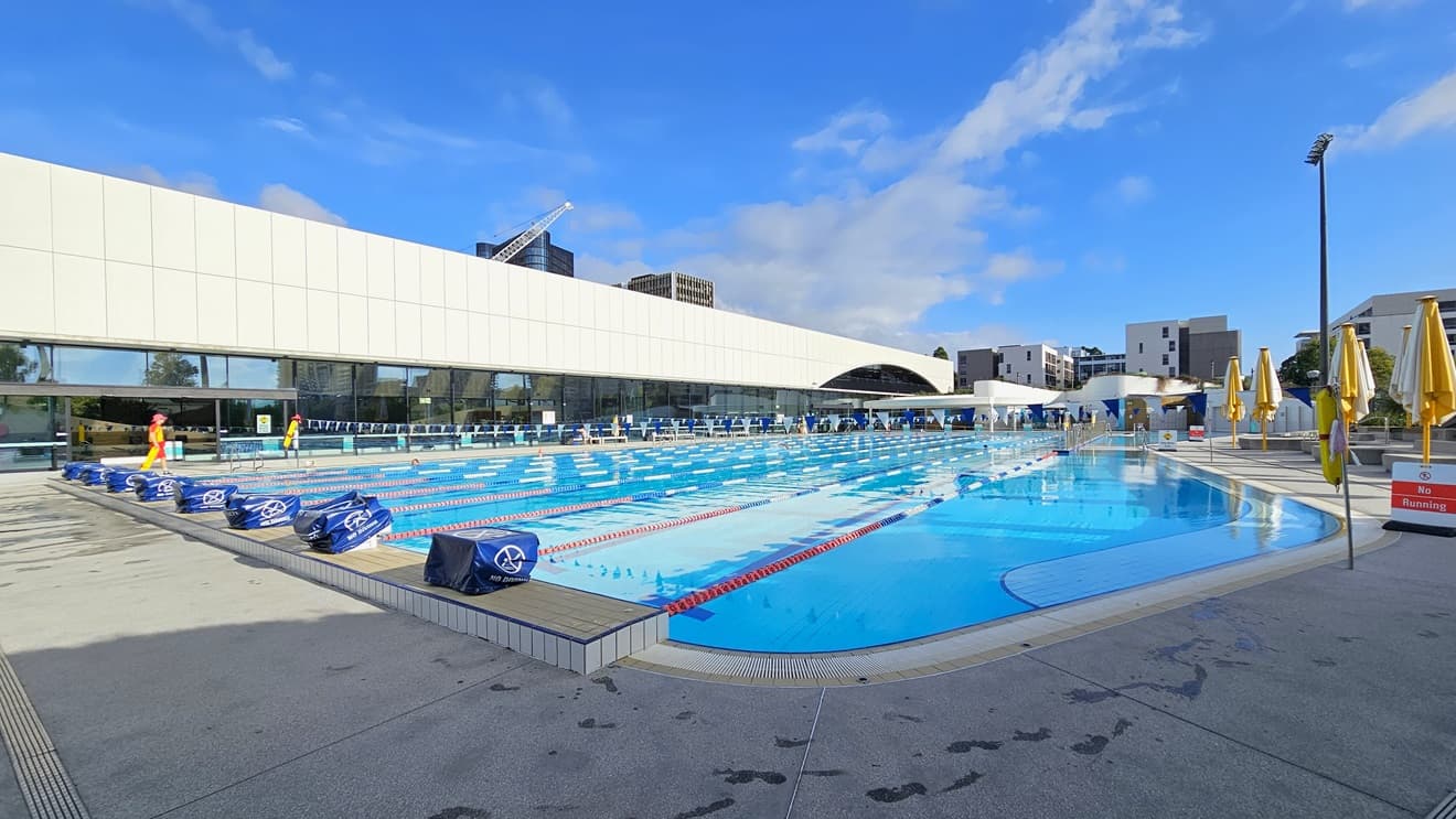 To the left is the facility building equipped with a white-tiled facade and glass walls. The 50m Pool is equipped with diving boards with covers. Safety banners and a hoist are positioned on the right near the pool overflow grid, along with folded umbrella shades. Colorful floating ropes and a string of triangle banners run across the pool. On the right, folded yellow umbrella shades. Tall floodlights and buildings are also visible.