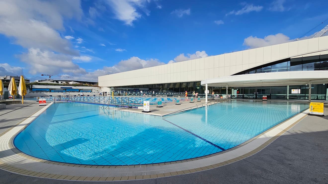 The Leisure Pool features stairs with a ramp entry and is surrounded by an overflow grid. On the right is a circular shade structure that offers shading to the Leisure Pool. The facility building is also adjacent. Adjacent to it is the 50m Pool equipped with floating ropes and triangle banners. In between these two pools is a pool deck equipped with safety banners. On the left, folded umbrella shades are also visible.