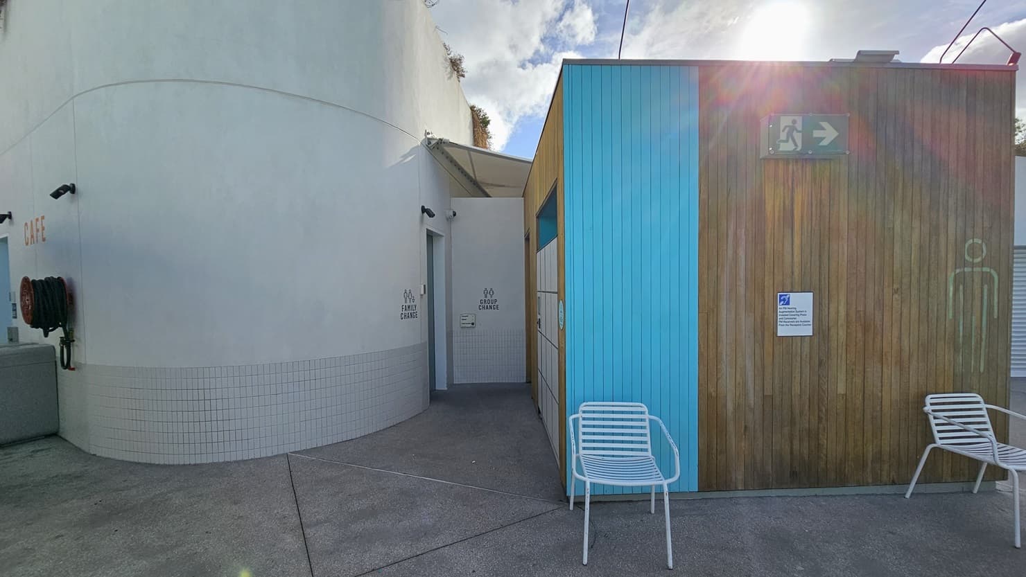 To the right is a Cabana-style Male and Female Change Room featuring a wooden design with a blue accent exterior. Signage is fixed to the wall. White chairs are positioned. To the left is a family change room featuring a concrete grey wall with small tiles accent. A fire hose reel is positioned on the left.