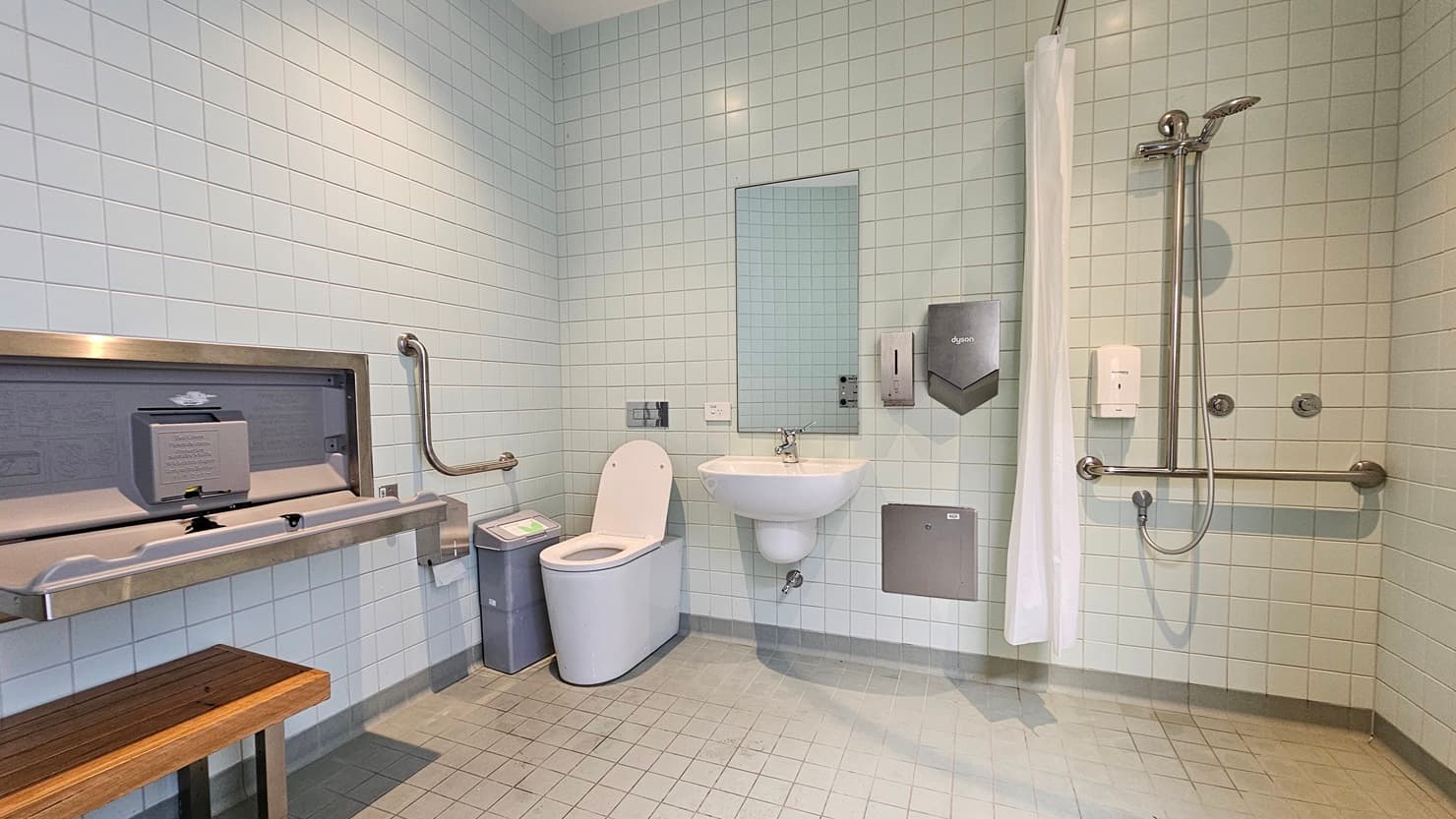 To the right is a shower equipped with handrails and a shower curtain. Next to it is the toilet bowl with handrails. A mirror, soap, and tissue dispenser are affixed to the wall. A rubbish bin and toilet brush are placed nearby. To the left is a wooden bench for seating and a foldable change table. The walls are covered with white tiles from floor to ceiling. The ceiling is white with LED lights, and the floor has white tiles.