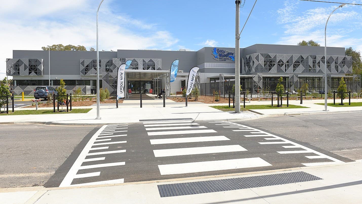 A modern building with geometric patterns on the facade is in the background. In front, there is a wide pedestrian crossing leading to the entrance. Several flags with the text "JOIN TODAY" are positioned near the entrance. The foreground features a paved road and a neatly maintained footpath with small trees and grass patches. The sky is partly cloudy.