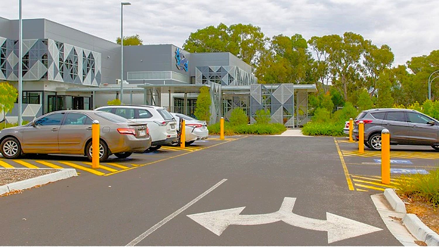 A modern building with geometric patterns on the facade is in the background, surrounded by trees. In the foreground, a car park with several vehicles, including a grey sedan and a silver SUV, is visible. Yellow bollards and painted lines mark the parking spaces. An arrow on the asphalt indicates the direction of traffic flow. The area is bordered by greenery and a kerb.