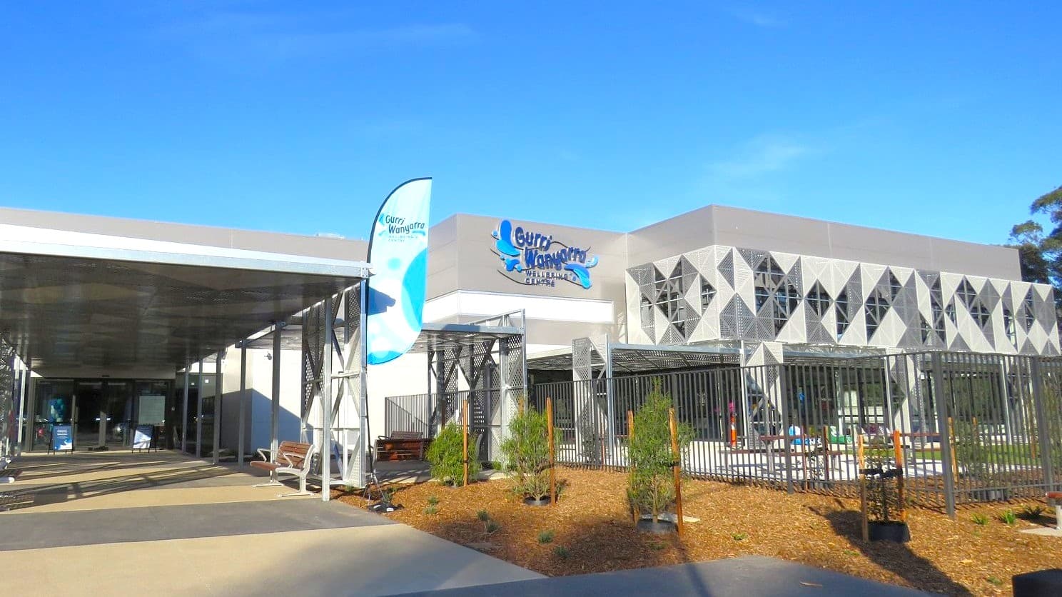 A modern building with a geometric facade featuring triangular patterns on the right. A sign with the name "Gurru Wanyarra" is visible. On the left, there's a covered walkway with metal supports and a blue flag. The foreground has a landscaped area with small plants and a metal fence. The ground is paved, and the sky is clear.