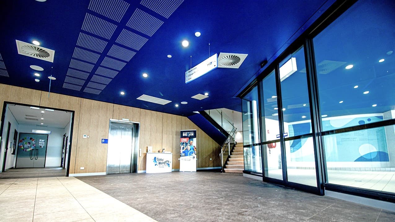 Three areas to explore Gurri Wanyarra Wellbeing Centre. Each section area of the venue can either be accessed by doors, an elevator, or a staircase. The walls of the entry points of each section are painted wooden brown, the ceilings are dark blue, and the tiles are grey to light brown.