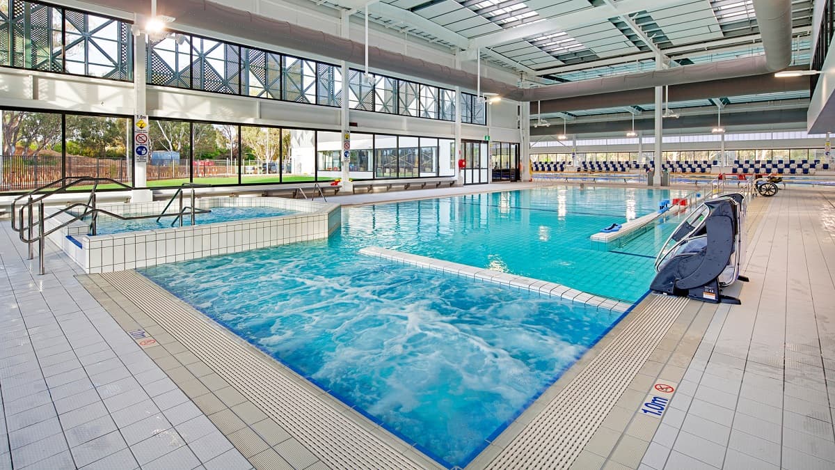 On the left, there's a section of the pool with a shallow area and grab rails for easy access. To the right, a lane for swimming extends towards the back, where swimming blocks are visible. The pool features clear blue water that reflects overhead lights. The flooring is tiled in a light colour, with drainage along the edges. The ceiling has a modern structure with large windows allowing natural light to flood in, enhancing the bright environment.