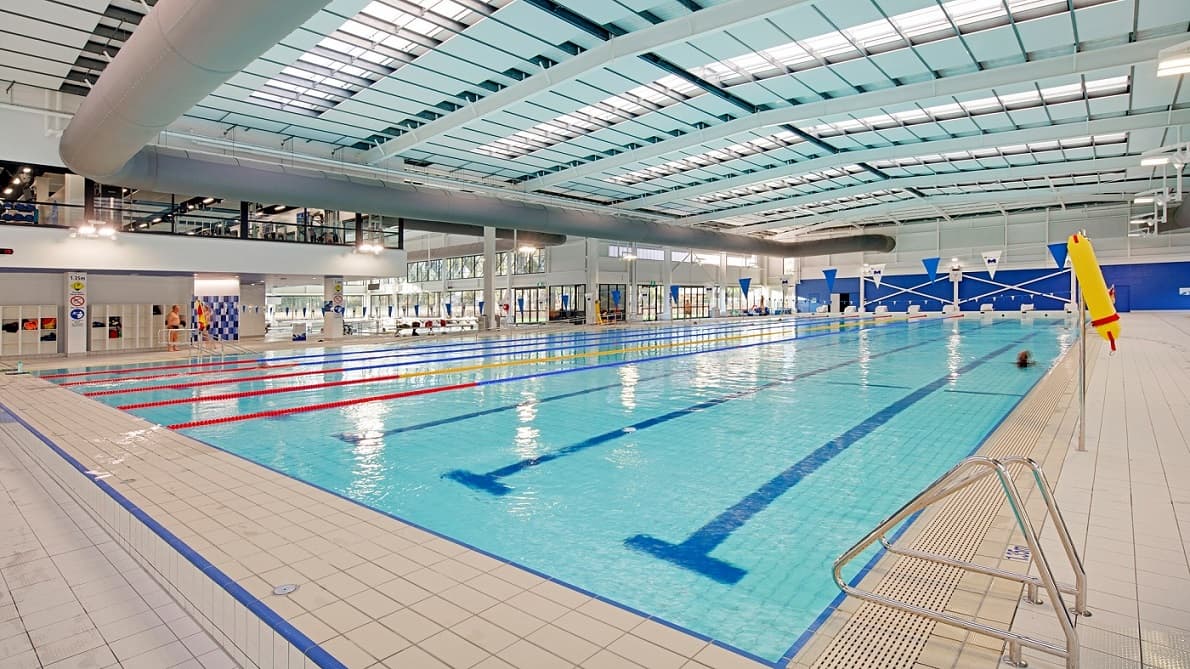On the left side, there's an area with lane markers and a swimming block positioned for use. Red and blue dividing lines streak across the clear water of the pool, which reflects bright overhead lights. To the right, a rescue buoy hangs, and additional lane markers are visible. The pool's flooring is tiled in a light colour, with drainage channels at the edges. The ceiling features a modern design with large windows that allow natural light.