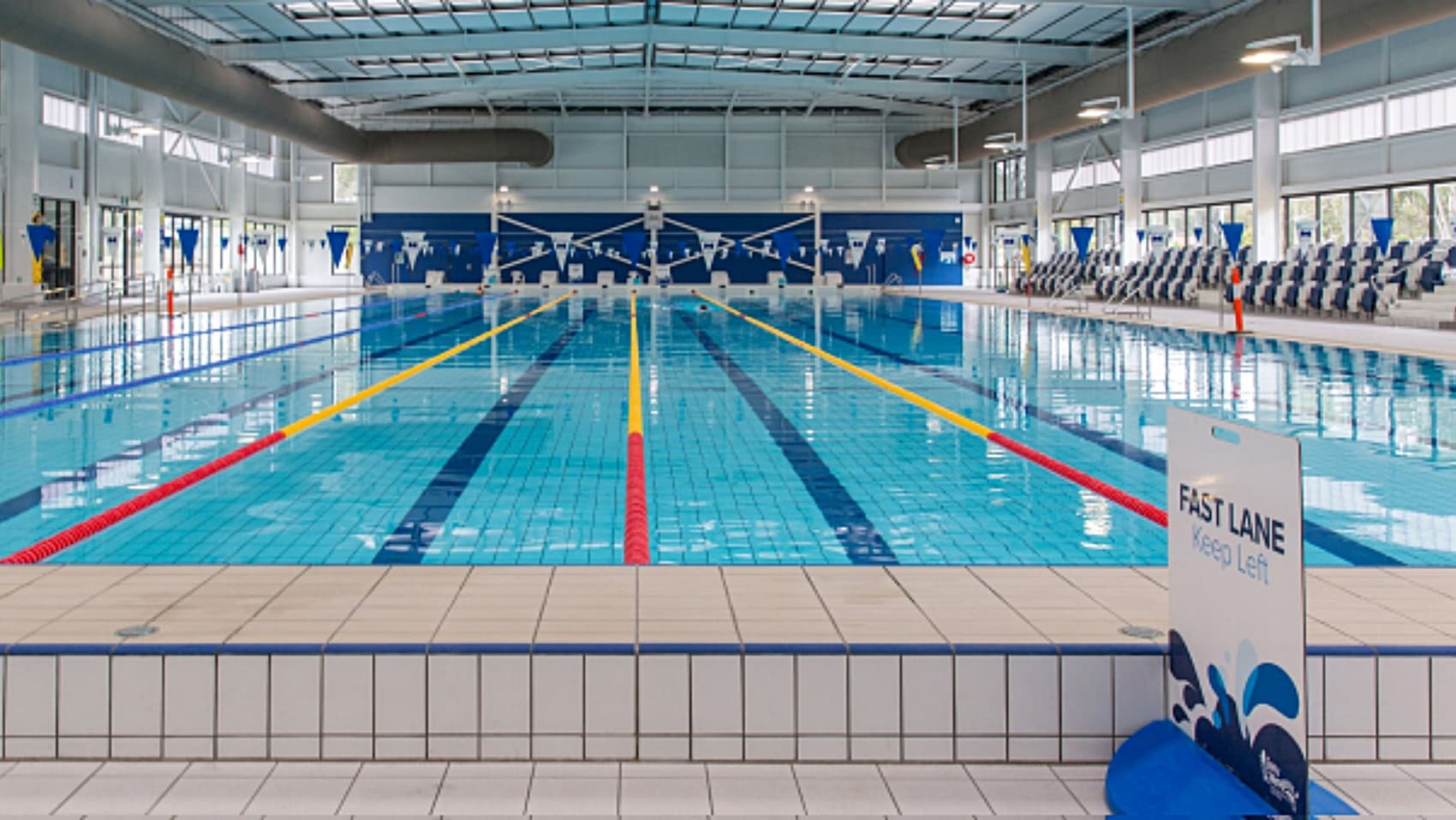 On the left, there's a white sign stating "FAST LANE Keep Left" situated on the pool edge. The clear water of the pool is marked with multiple lane lines in red, yellow, and blue. In the centre, several lanes are visible, each with distinct colours guiding swimmers. To the right, there are rows of poolside seating equipped with grey chairs. Large windows in the background allow natural light to enter, illuminating the area.