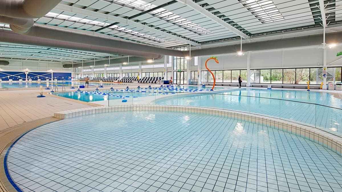 To the left, there's a rounded section of the pool with light blue tiles leading into the deeper area. A bright orange water slide curves upwards, adding a playful element. In the centre, several lanes are marked, and swim blocks are visible. On the right, a section of pool furniture is set up with chairs against the wall, which features large windows allowing natural light to enter. The flooring is tiled and light, surrounded by drainage channels.