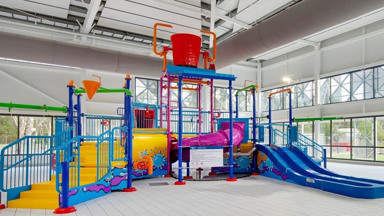 In the centre, there's a colourful water play structure featuring multiple slides and climbing areas. A large red bucket sits at the top, poised to tip water down, while a purple slide curves down to the right. Adjacent to it, a small orange funnel hangs, with bright green pipes extending outwards. The floor is tiled in a light colour, providing a clean surface beneath the play area. The ceiling is high with exposed ductwork and ample lighting, enhancing the spacious feel of the environment.