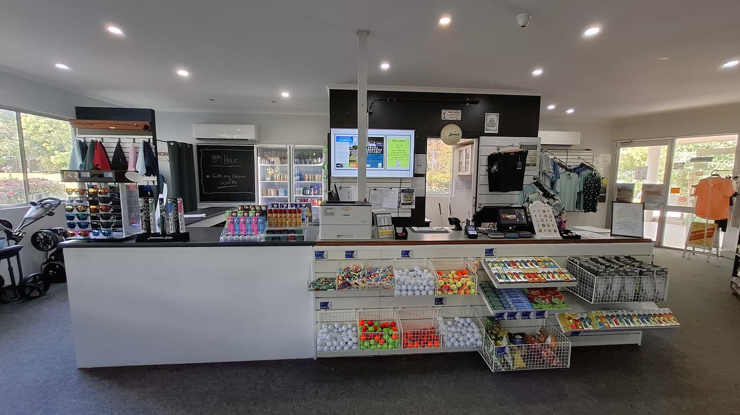On the left, a display rack holds various coloured caps and sunglasses, with a golf trolley positioned nearby. The floor is covered in dark carpet, leading to a service counter in the centre. The counter features a cash register and a selection of snacks and golf balls in clear containers. On the right, a large glass door opens to the outside, with shirts hanging on a rack nearby. The ceiling is flat and white, with recessed lighting.