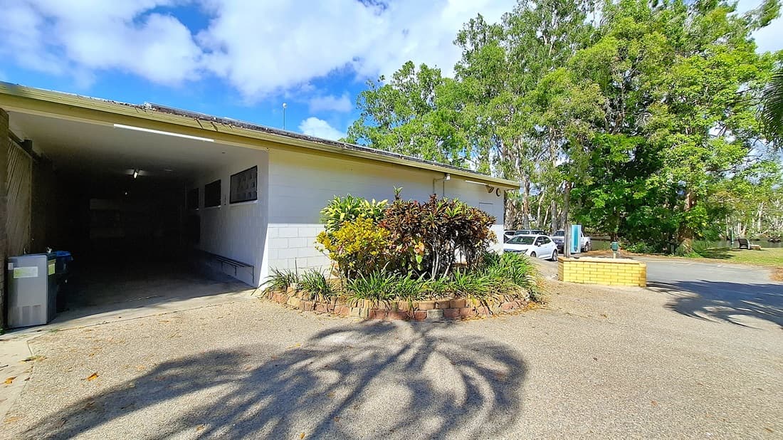 On the left, a garage entrance is visible, with a concrete floor and a few fluorescent lights illuminating the interior. The wall beside the garage is painted white, featuring several small windows. In the centre, a circular garden bed is surrounded by a low brick border, filled with various plants and shrubs. On the right, a white car is parked next to the garden, with a pathway leading further into the area. The ground is a mix of concrete and gravel.