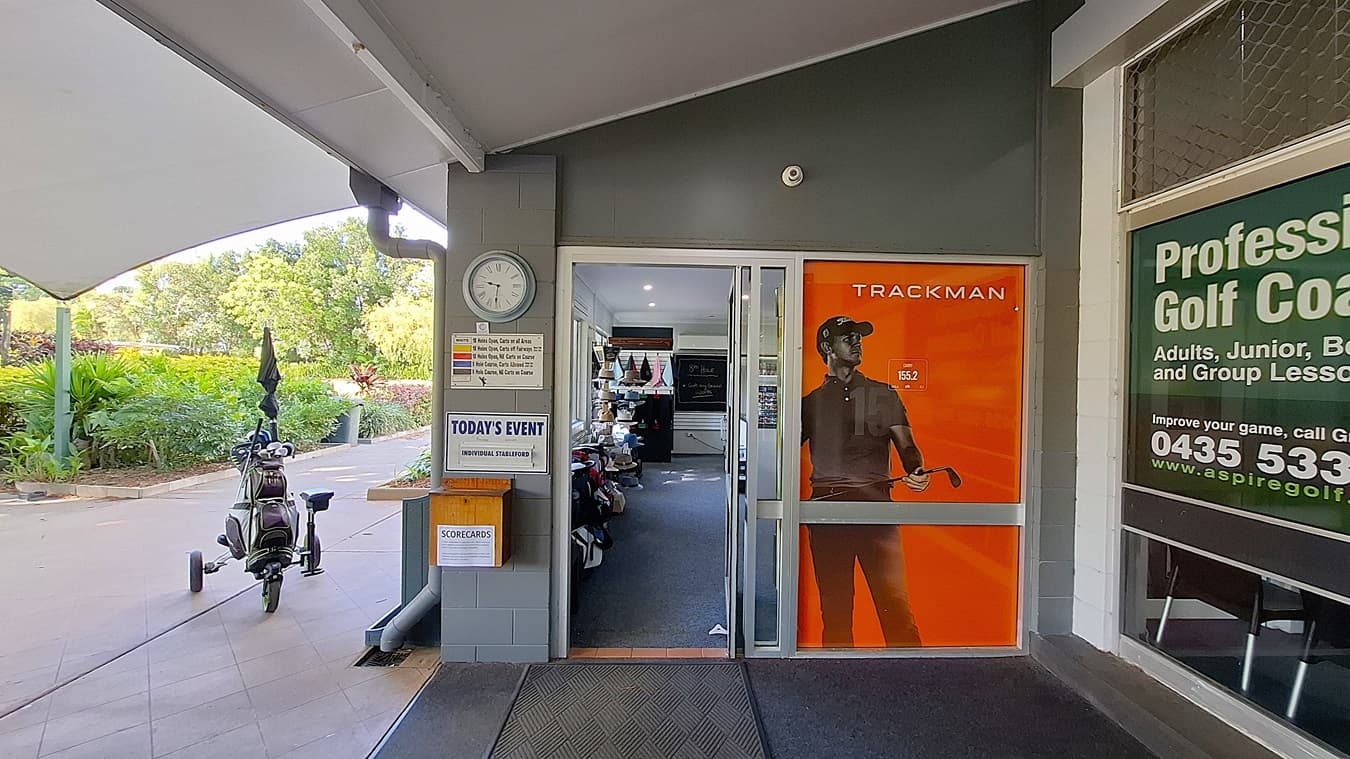 On the left, a clock is mounted on the wall beside a notice board displaying event information. A golf bag on wheels is parked nearby, with an umbrella attached. The entrance features a glass door with an orange advertisement for "TRACKMAN." In the centre, the floor is covered with dark carpet leading into the shop area, which has shelves filled with golf equipment. On the right, a large window displays information about golf coaching services. The ceiling is flat and white.