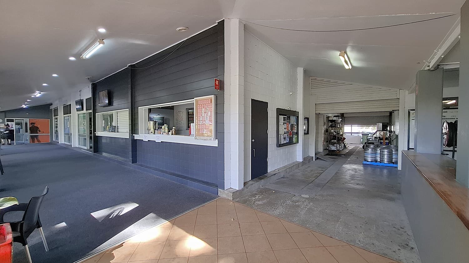 On the left, a long corridor features a dark carpeted floor, with several windows and a service counter displaying drinks and snacks. The walls are painted in light colours, with a few televisions mounted above. In the centre, the corridor leads to an open area with a tiled floor, transitioning to a concrete surface. On the right, a large opening reveals a storage area with kegs and equipment. The ceiling is flat and painted white, with fluorescent lights.