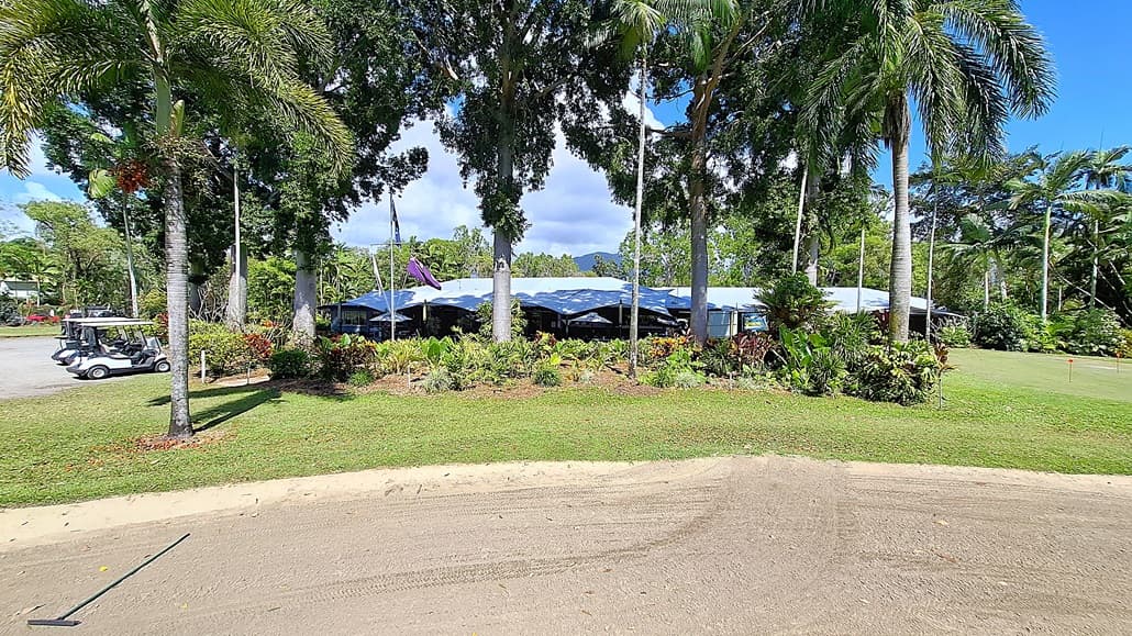 On the left, a row of golf carts is parked on a gravel surface, with a few palm trees providing shade. The ground is a mix of grass and sandy areas, leading towards a landscaped garden filled with various plants. In the centre, a building with a blue roof is partially obscured by trees, featuring large windows and outdoor seating areas. On the right, more greenery and trees create a natural border, enhancing the outdoor setting.