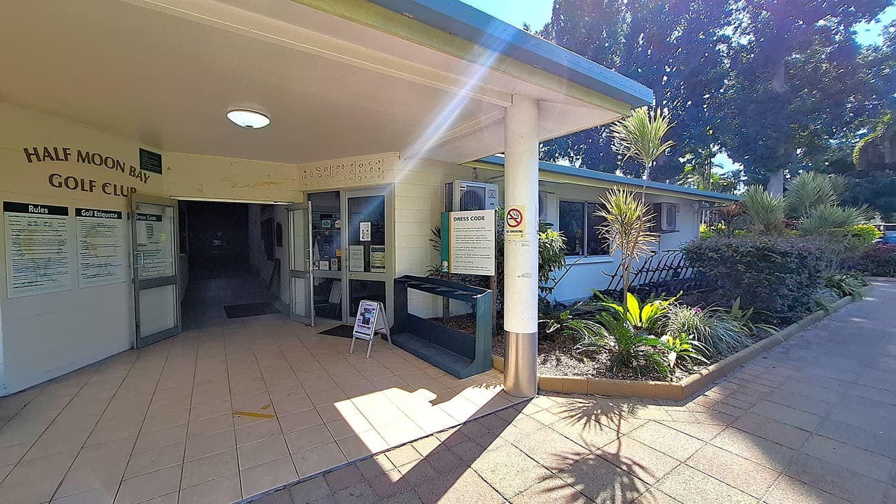 On the left, the entrance to the Half Moon Bay Golf Club features double glass doors, with a tiled floor leading inside. A notice board displays rules and etiquette for visitors. In the centre, the corridor extends into a darker area, while a service counter is visible to the right, with a sign detailing the dress code. On the right, a landscaped area includes various plants and a bench. The ceiling is flat and white, with a circular light fixture.