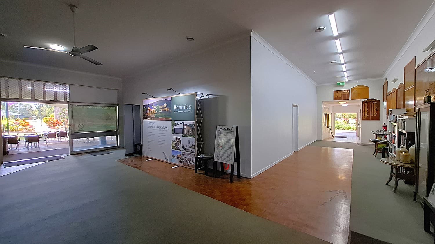 On the left, a large display stand features promotional materials for a local business, with a small table beside it. The floor is a mix of light-coloured tiles and a darker wooden section. In the centre, the corridor leads towards a doorway, with a light grey carpet underfoot. On the right, a wall showcases wooden plaques and a small shelf with items for sale. The ceiling is flat and white, with fluorescent lights.