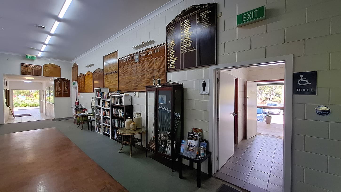 On the left, a bookshelf holds various books and pamphlets, with a small table displaying two large containers. The floor is a combination of light tiles and a darker carpet. In the centre, a corridor leads to an exit, with large wooden boards mounted on the wall displaying names and scores. On the right, a doorway leads to the toilets, marked with signs for "Gentlemen" and "Toilet." The ceiling is flat and white, with fluorescent lights.