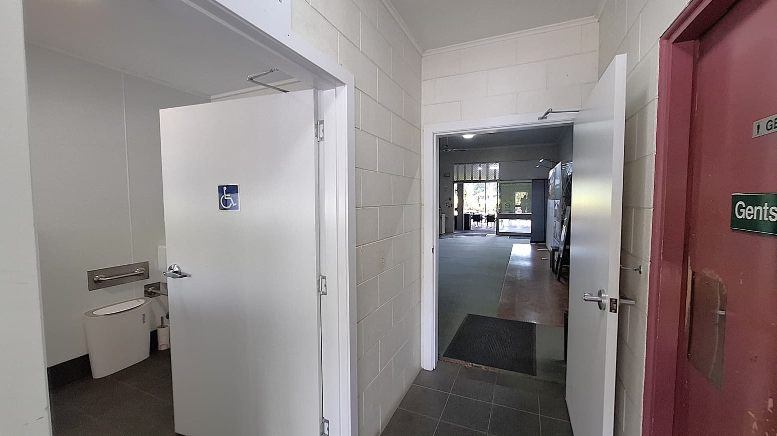 On the left, a white door leads to a disabled toilet, with a visible toilet and grab bars inside. The floor is dark tiled, contrasting with the light-coloured walls. In the centre, a corridor extends towards a larger room, with a smooth grey floor and a doorway leading outside. On the right, a red door is marked "Gents," indicating the men's toilet. The ceiling is flat and white, with overhead lights illuminating the area.