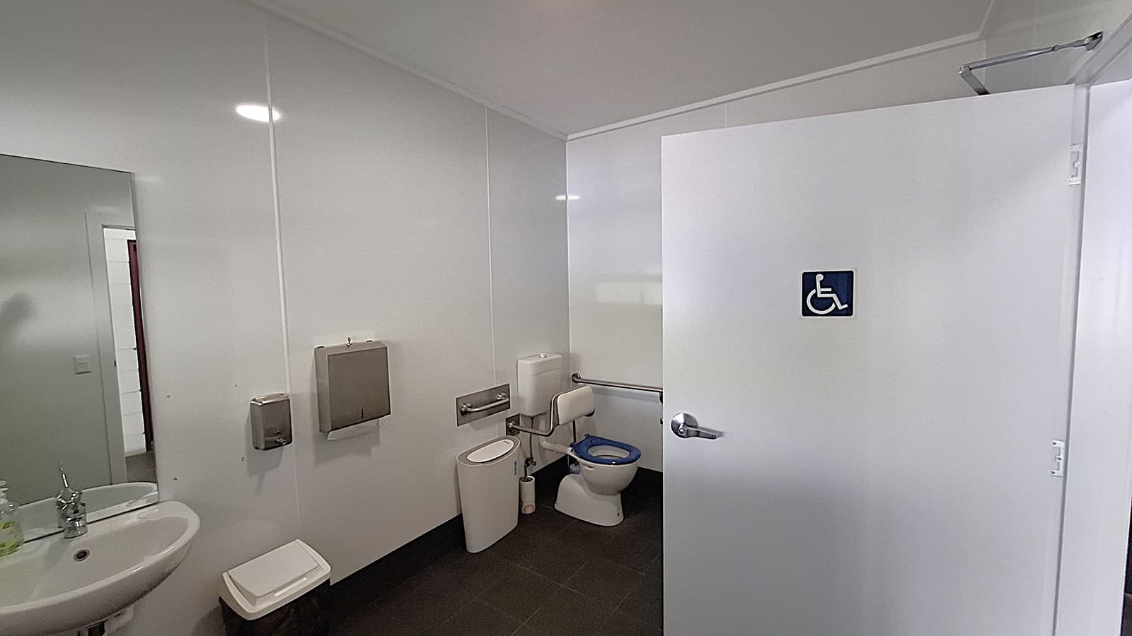On the left, a white sink with a chrome tap is mounted on the wall, next to a small bin. Above the sink, a mirror reflects the space. The floor is dark tiled, providing a contrast to the light-coloured walls. In the centre, a toilet with a blue seat is positioned beside a wall-mounted grab rail. On the right, a white door marked with a blue accessibility symbol leads to a disabled toilet. The ceiling is flat and white, with overhead lights illuminating the area.