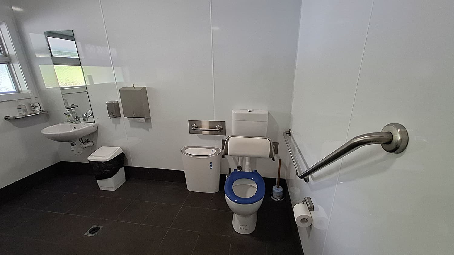 On the left, a white sink is mounted on the wall, with a mirror above it reflecting the space. A small shelf holds toiletries, and a bin is positioned below the sink. The floor is dark tiled, providing a contrast to the light walls. In the centre, a toilet with a blue seat is equipped with grab bars for accessibility. On the right, a roll of toilet paper is mounted on the wall, next to a silver handrail. The ceiling is flat and white, with overhead lights.