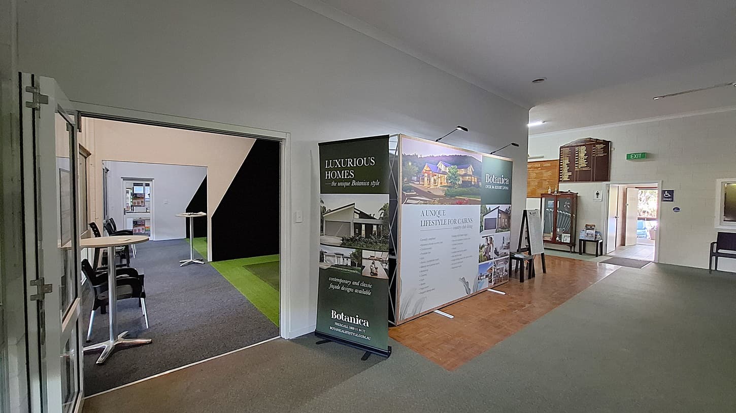 On the left, a large display stand features promotional materials for luxurious homes, with images and text highlighting various properties. A small table with two chairs is positioned nearby. The floor is a mix of dark carpet and wooden tiles. In the centre, a corridor leads to another room, with a green carpet visible on the right side. On the right, a wooden board lists names and achievements, and a doorway leads to a restroom. The ceiling is flat and white, with overhead lights.