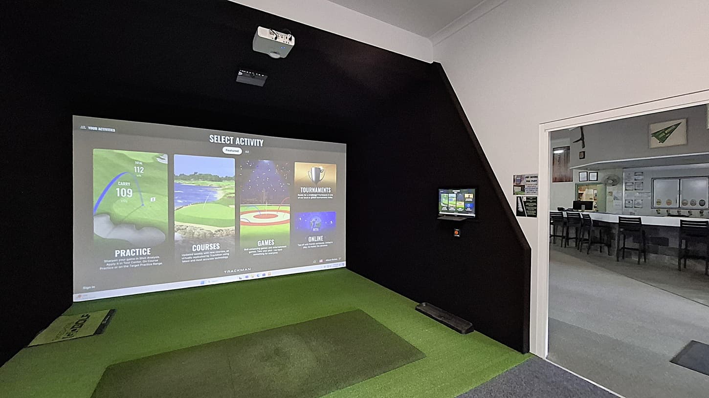 On the left, a projection screen displays options for golf activities, including practice and courses, with a green mat positioned in front for play. The floor is covered in artificial grass. In the centre, the space is enclosed by black walls, creating a focused environment. On the right, a small monitor shows additional information, and a doorway leads to another room with tables and chairs visible. The ceiling is flat and white, with a projector mounted above.