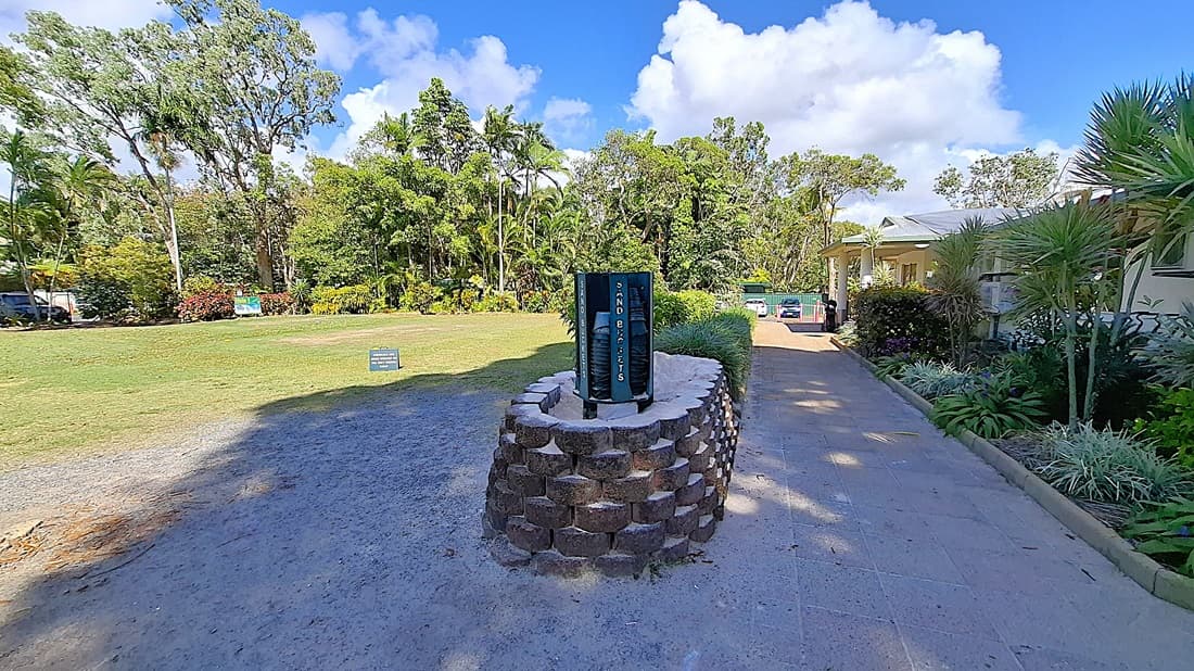 On the left, a grassy area is bordered by trees and shrubs, with a small sign indicating "Sand Tickets" positioned on the ground. The pathway is made of light-coloured tiles, leading towards a building on the right. In the centre, a circular stone feature stands with a vertical sign displaying information. On the right, the building has a light-coloured exterior and is surrounded by tropical plants.