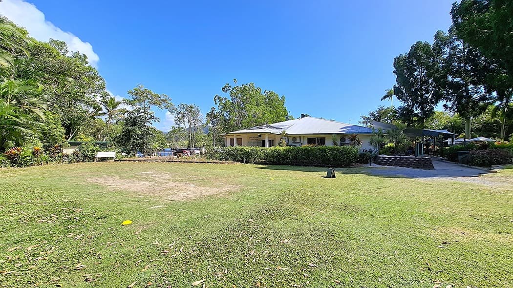 On the left, a grassy area is bordered by a variety of trees and shrubs, with a bench positioned for seating. The ground is mostly green, with some patches of bare earth. In the centre, a single-storey building features a light-coloured exterior and several air conditioning units mounted on the walls. On the right, a covered outdoor seating area is visible, surrounded by more greenery. The floor is a mix of grass and gravel.