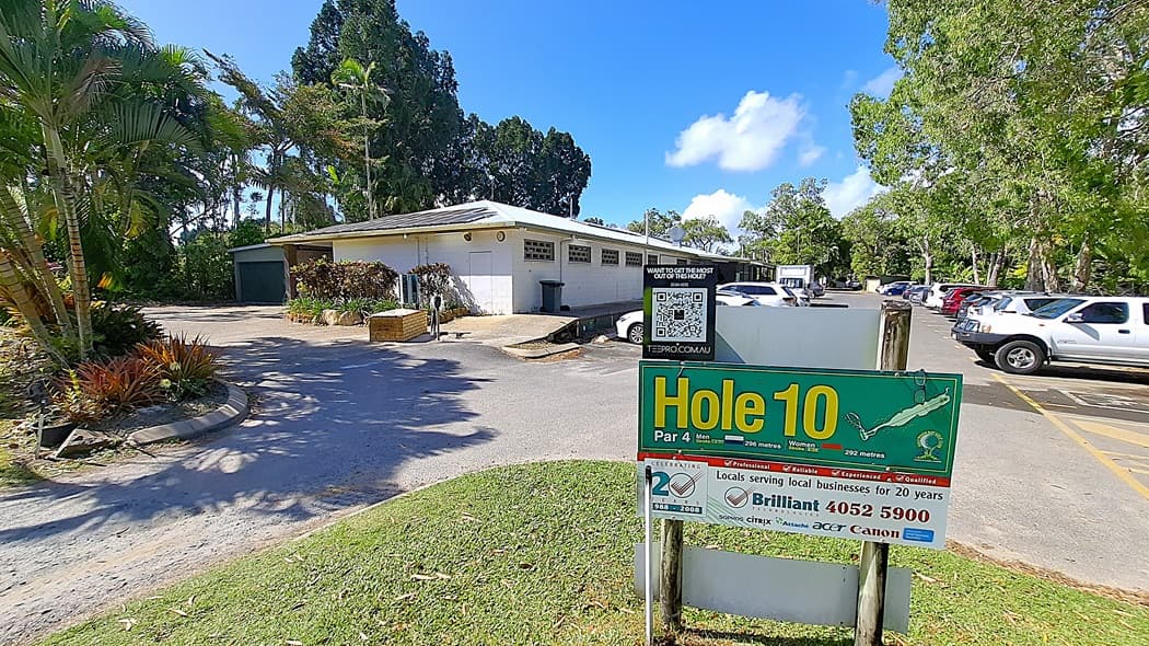 On the left, a building with a light-coloured exterior features several windows and a door, with a small area of landscaping including shrubs and a palm tree. A bin is positioned next to the building. In the centre, a sign reading "Hole 10" is prominently displayed, indicating the golf course hole details. On the right, a parking area is visible with multiple vehicles parked in a gravel lot. The ground is a mix of gravel and grass.