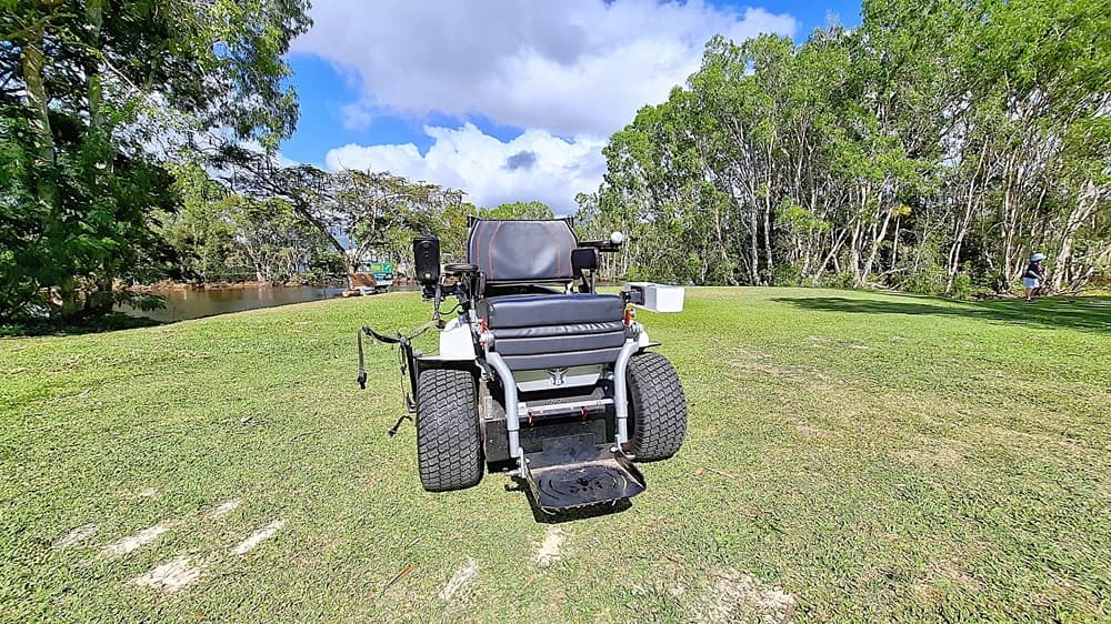 On the left, a patch of grass is visible, bordered by trees that provide shade. A small body of water can be seen in the background, reflecting the greenery. In the centre, a golf cart is parked, featuring a black seat and large wheels, with controls visible on the sides. On the right, a person is walking in the distance, partially obscured by trees. The ground is a well-maintained lawn.