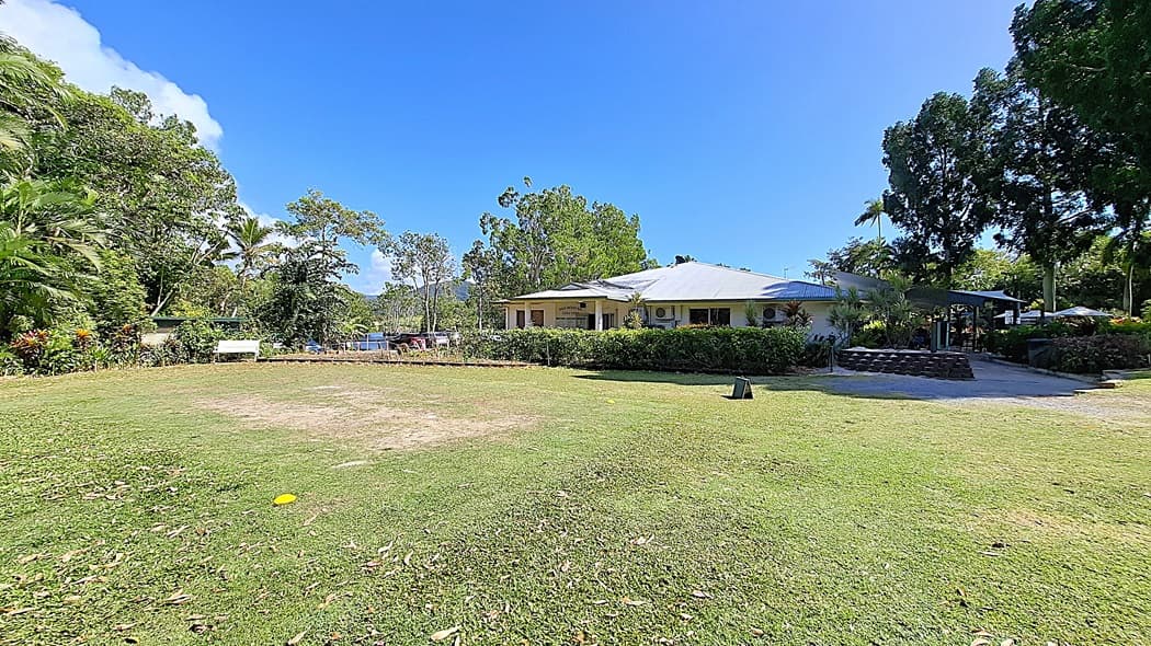 On the left, a grassy area is bordered by a variety of trees and shrubs, with a white bench positioned for seating. In the centre, a single-storey building features a light-coloured exterior and several air conditioning units mounted on the walls. On the right, a covered outdoor area is visible, surrounded by more greenery and a stone pathway leading to it. The ground is mostly grass with some bare patches.