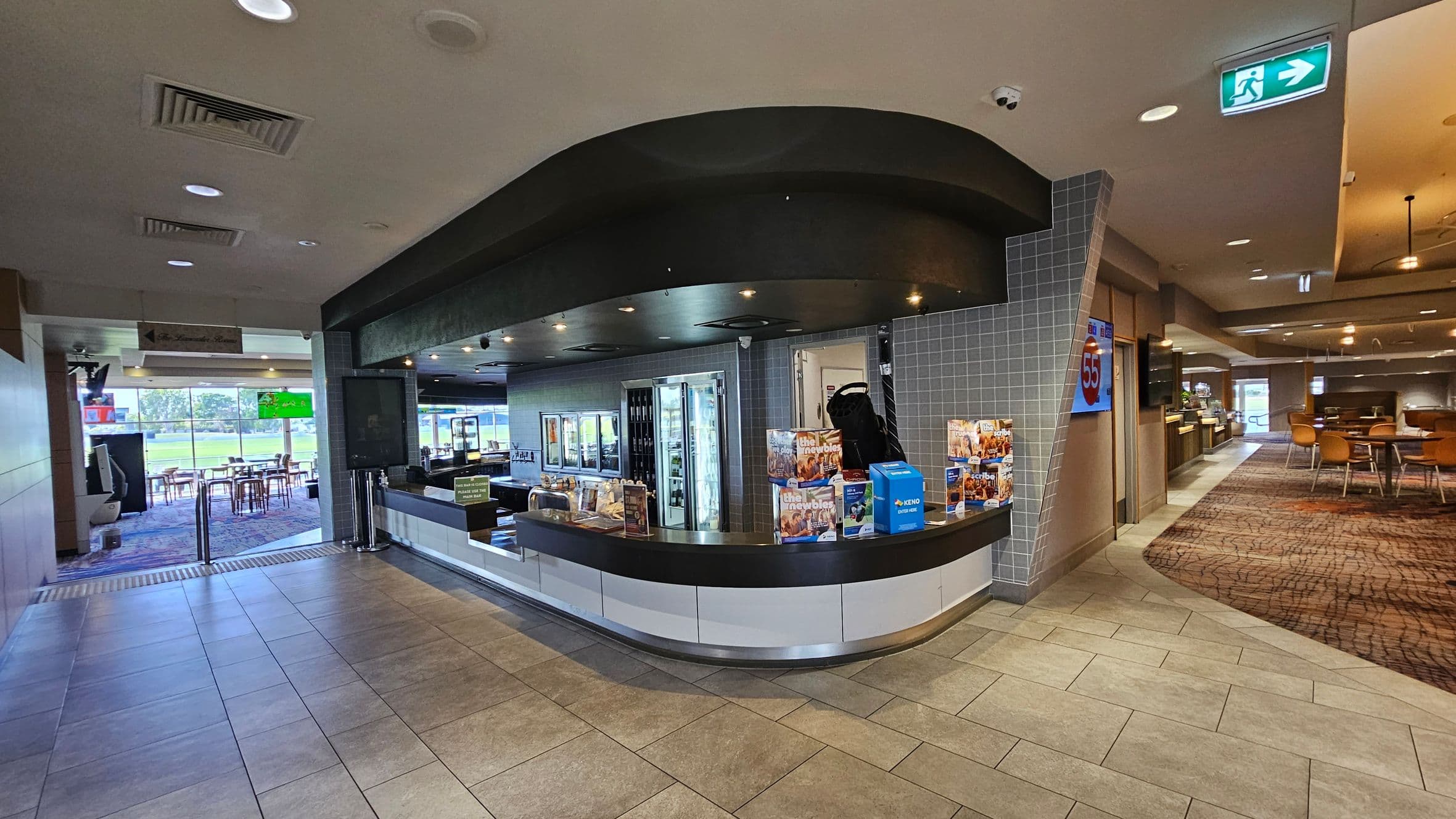 A spacious dining area with numerous wooden tables and chairs arranged in rows. The floor is covered with a patterned carpet in shades of blue and red. On the left, there are more tables and a menu stand with the number 29. The ceiling is high with large, woven light fixtures hanging down. In the background, a sign reads "The Lunch Room" on a wall, and there are potted plants near the entrance. The ceiling has recessed lighting and air vents.