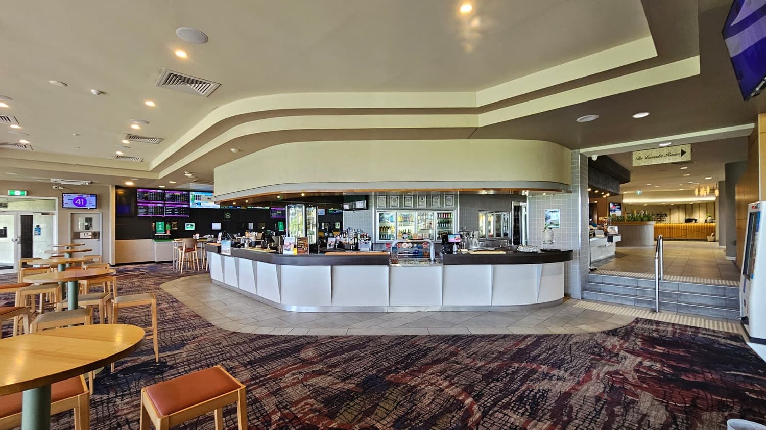 A spacious interior with a modern bar in the centre, featuring a curved counter and a variety of drinks displayed behind. On the left, there are several wooden tables and chairs on a patterned carpet. The ceiling is adorned with recessed lighting. To the right, a tiled floor leads up a few steps to another area with more seating and greenery. Multiple screens displaying information are mounted on the walls.
