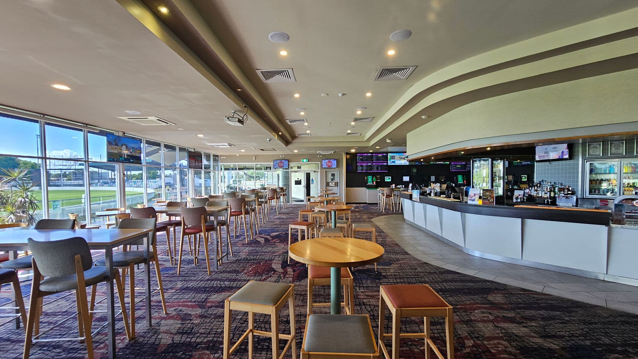 A spacious indoor area with a modern bar on the right, featuring a curved counter and a variety of drinks displayed. On the left, large windows provide a view of a green outdoor area, with several flat-screen TVs mounted above. The centre has high tables and chairs on a patterned carpet. The ceiling is equipped with recessed lighting and air conditioning vents.