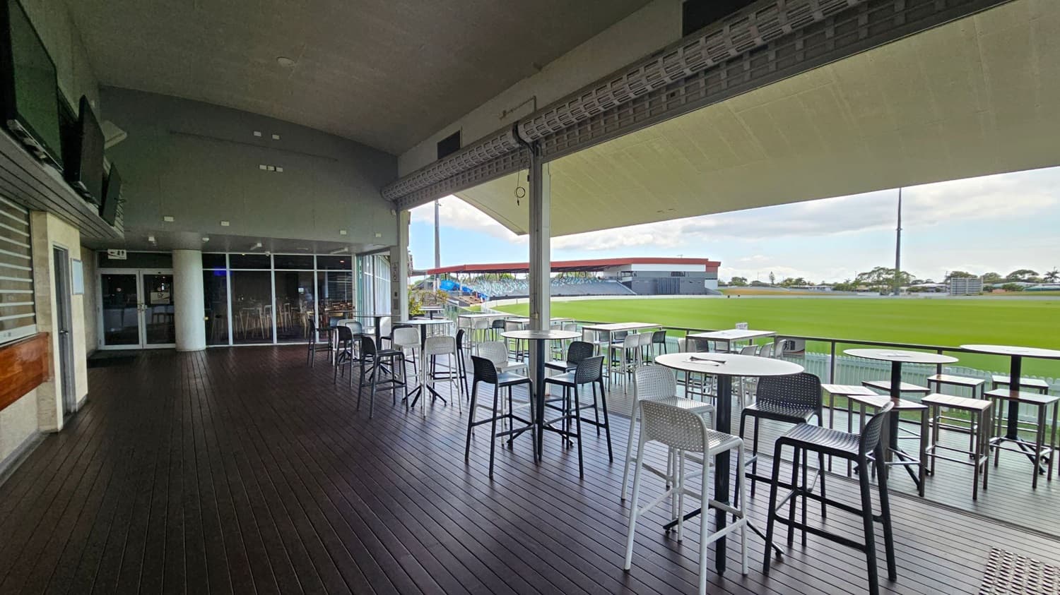 A spacious outdoor seating area with dark wooden flooring and a high ceiling. On the left, there are several wall-mounted televisions and a glass door leading inside. In the middle, there are multiple high tables with stools, some in white and others in black. On the right, the area opens up to a view of a green sports field with a distant grandstand. The ceiling is light-coloured, and the area is partially covered, providing shade.