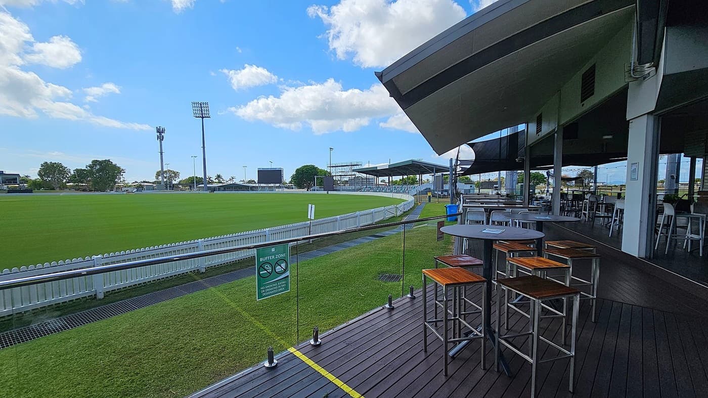 A cricket ground with a lush green field is on the left, surrounded by a white picket fence. Tall floodlights and a scoreboard are visible in the background. On the right, there is a covered seating area with high tables and wooden stools on a wooden deck. A glass barrier separates the seating area from the field, with a sign indicating a buffer zone. The sky is partly cloudy.