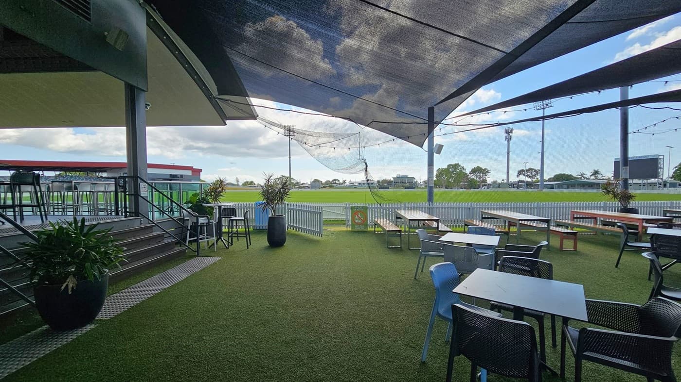 Outdoor seating area with artificial grass flooring. On the left, there are steps leading to a raised platform with high stools and tables. In the centre, potted plants and a white picket fence border a sports field. On the right, several tables and chairs are arranged under a canopy. Overhead, string lights are visible. In the background, a large scoreboard and floodlights can be seen, along with trees and a distant building.