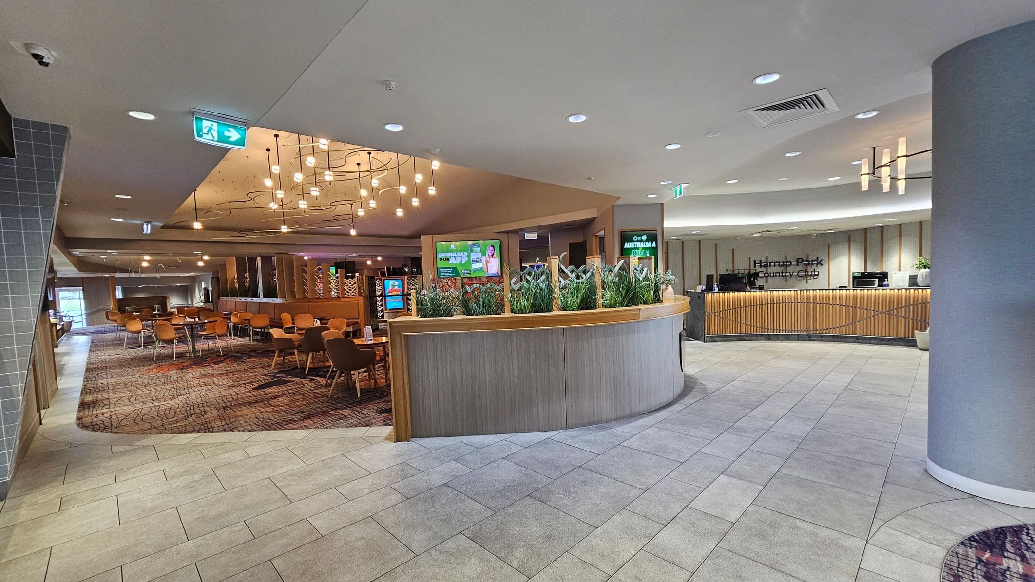 A modern interior with a curved reception desk on the right, featuring a sign for Harrup Park Country Club. Behind the desk are vertical wooden slats and potted plants. In the centre, a partition with greenery separates the space. On the left, a dining area with wooden tables and chairs on a patterned carpet. The ceiling has recessed lighting and decorative hanging lights. The floor is tiled in light grey.