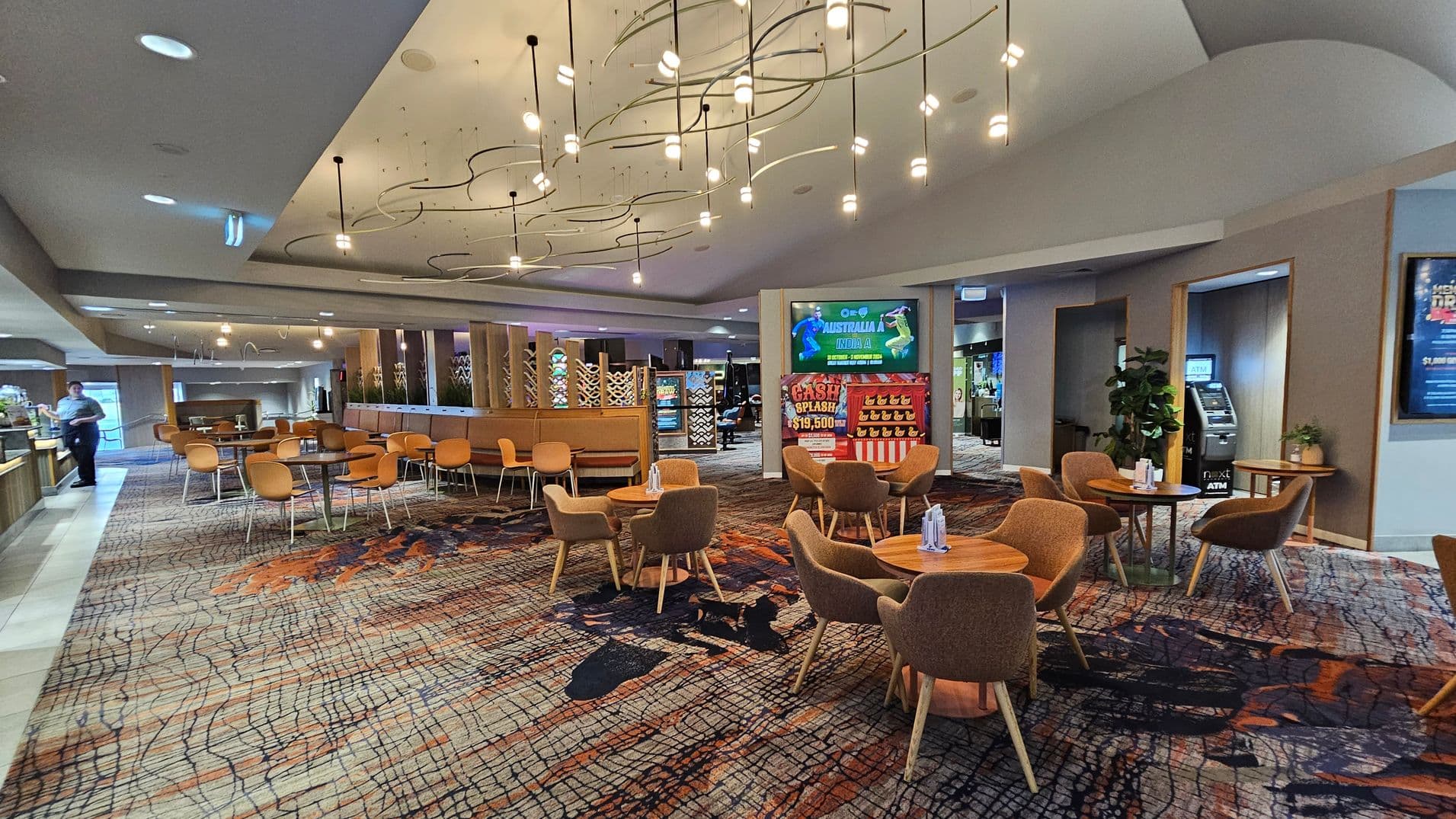 A spacious indoor area with a patterned carpet in shades of orange, black, and beige. On the left, there are several round tables with orange chairs, and a person is visible near a counter. In the centre, more tables with brown chairs are arranged. The ceiling features modern, artistic light fixtures. On the right, a digital screen displays a sports event, and an ATM is situated nearby. The walls have decorative panels and plants.
