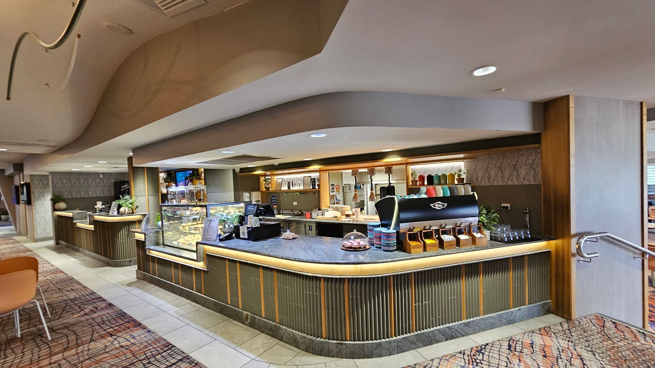 A modern café counter with a marble top is in the centre, featuring a coffee machine with colourful cups stacked on top. To the left, a glass display case shows various pastries. Behind the counter, shelves hold more cups and decorative plants. The floor is a mix of tiled and patterned carpet, and the ceiling has a curved design with recessed lighting. On the right, a metal handrail is visible, leading to a seating area with wooden chairs.